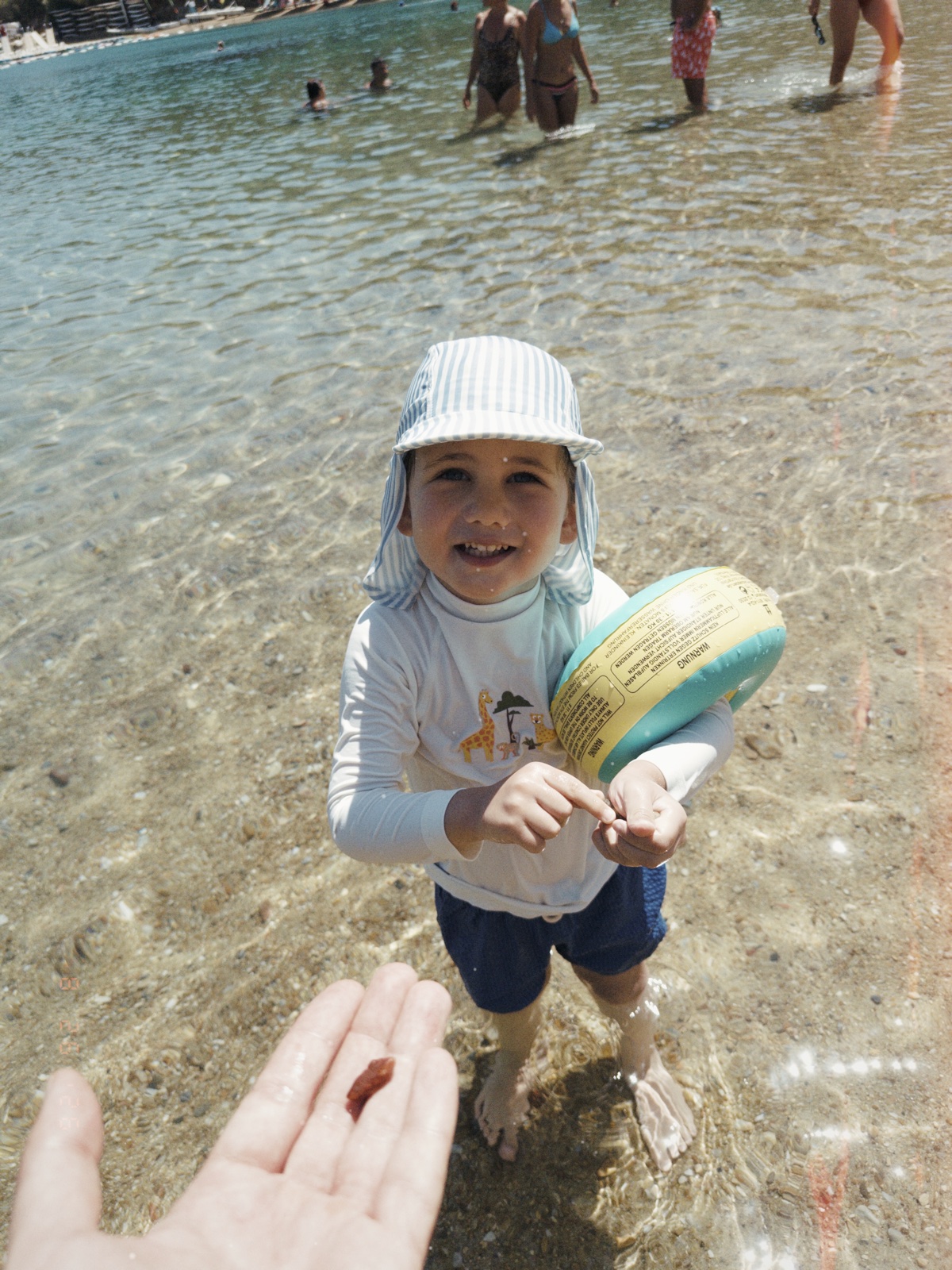 A child with a striped sun hat and swimming float standing in the sea.